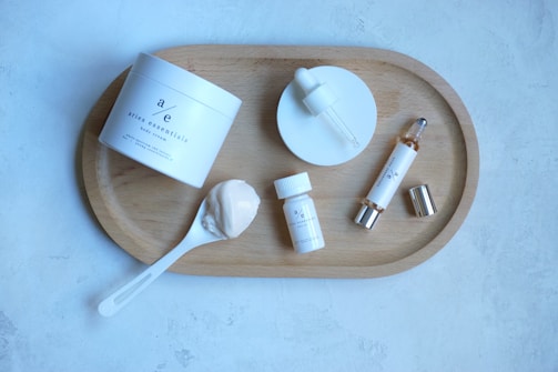 A neatly arranged skincare set featuring various products on a wooden tray. Included are a jar of body cream, a dropper bottle, a small bottle, a spoon with cream, and a cylindrical bottle with a silver cap. The background is a light gray textured surface.