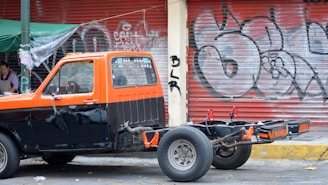 A parked pickup truck missing its bed with a two-tone paint job of orange and black is captured beside a street vendor under a green tarp. In the background, a red metal shutter door is covered with graffiti.