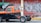 A parked pickup truck missing its bed with a two-tone paint job of orange and black is captured beside a street vendor under a green tarp. In the background, a red metal shutter door is covered with graffiti.