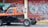 A parked pickup truck missing its bed with a two-tone paint job of orange and black is captured beside a street vendor under a green tarp. In the background, a red metal shutter door is covered with graffiti.