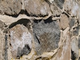 Close-up of a textured stone wall composed of irregularly shaped rocks. The stones are of varying shades of gray and brown, with some white patches. Sand-colored mortar fills the gaps between the stones, providing a rough and rugged appearance.
