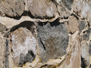 Close-up of a textured stone wall composed of irregularly shaped rocks. The stones are of varying shades of gray and brown, with some white patches. Sand-colored mortar fills the gaps between the stones, providing a rough and rugged appearance.