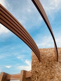 Curved metal beams stretch across a clear blue sky, supported by sturdy stone walls constructed from irregular, warm-colored bricks. The scene conveys a blend of modern architectural design with natural materials.