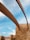 Curved metal beams stretch across a clear blue sky, supported by sturdy stone walls constructed from irregular, warm-colored bricks. The scene conveys a blend of modern architectural design with natural materials.