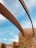 Curved metal beams stretch across a clear blue sky, supported by sturdy stone walls constructed from irregular, warm-colored bricks. The scene conveys a blend of modern architectural design with natural materials.