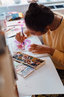 A person engrossed in painting with watercolors, using a vibrant palette and brush to create floral designs on paper. The workspace is filled with artistic tools and materials, creating a creative atmosphere.
