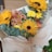 A bouquet of vibrant flowers including sunflowers, daisies, and chrysanthemums is wrapped in cream-colored paper. The arrangement features yellow, orange, and pink blooms with green eucalyptus leaves adding contrast.