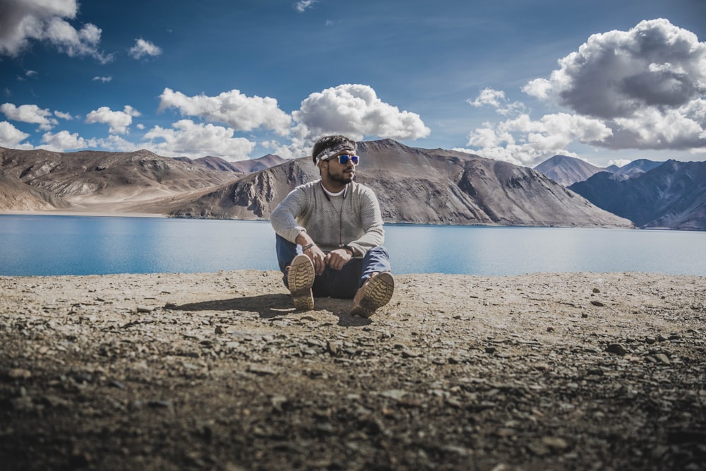 man in gray and white striped sweater sitting on brown rock near lake during daytime