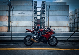 A red and black motorcycle is parked on a road in front of a tall stack of shipping containers, surrounded by a chain-link fence. The containers are industrial and feature branding. The overcast sky adds a moody atmosphere to the scene.