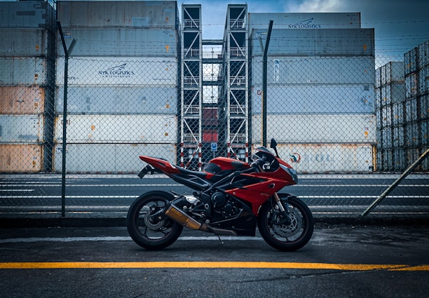 A red and black motorcycle is parked on a road in front of a tall stack of shipping containers, surrounded by a chain-link fence. The containers are industrial and feature branding. The overcast sky adds a moody atmosphere to the scene.