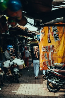 A busy street market scene with people walking through a narrow alley lined with small shops. A person wearing a blue helmet is sitting on a motorbike loaded with bags. Another person is walking away, dressed in casual clothing and a headscarf. A large banner advertising a beverage is prominently displayed on the right, with various goods and motorbikes visible along the sides.