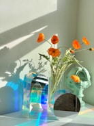 A collection of objects is arranged on a white surface, featuring vibrant orange flowers in a glass vase. Next to the vase is a decorative bust with one flower tucked into it. There are also geometric glass structures reflecting a spectrum of colors onto the surface and wall, under soft natural lighting.