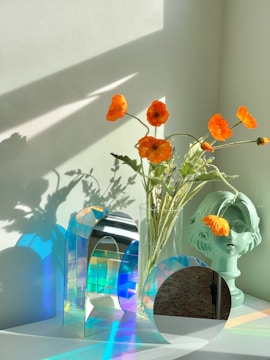 A collection of objects is arranged on a white surface, featuring vibrant orange flowers in a glass vase. Next to the vase is a decorative bust with one flower tucked into it. There are also geometric glass structures reflecting a spectrum of colors onto the surface and wall, under soft natural lighting.