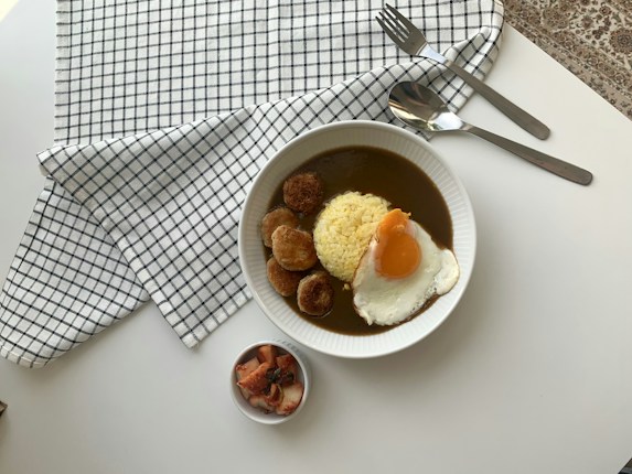 A bowl contains rice topped with a sunny-side-up egg, surrounded by a bed of curry and several fried items. The setting includes a small dish with pickled vegetables, placed on a white table covered with a black-and-white checkered cloth. To the side, there is a fork and spoon laid neatly.