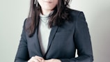 A woman with long dark hair is wearing a dark blazer over a light-colored turtleneck. She has her hands folded and is adorned with long dangling earrings. The background is a soft, neutral color.