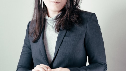 A woman with long dark hair is wearing a dark blazer over a light-colored turtleneck. She has her hands folded and is adorned with long dangling earrings. The background is a soft, neutral color.