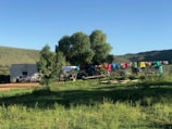 A rural landscape with a small white building and a large tree in the background. There is a line of colorful clothes hanging to dry. The surroundings are grassy, with a dirt pathway and rolling hills under a bright blue sky.