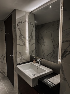 A modern bathroom with marble-patterned tiles covering the walls and sink area. The space features a large mirror, an inset sink with a chrome faucet, and a bottle of soap or lotion on the countertop. Soft overhead lighting creates a sleek and clean ambiance.