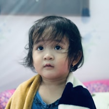 A toddler with big eyes and short hair looks forward with a thoughtful expression. The child is wrapped in a striped blanket and is wearing a blue shirt.