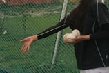 A person dressed in dark sportswear holds multiple baseballs in one hand while reaching out with the other hand. The background consists of a green mesh net and grassy area, suggesting a sports field or training area.