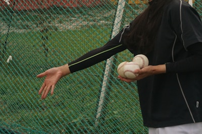 A person dressed in dark sportswear holds multiple baseballs in one hand while reaching out with the other hand. The background consists of a green mesh net and grassy area, suggesting a sports field or training area.