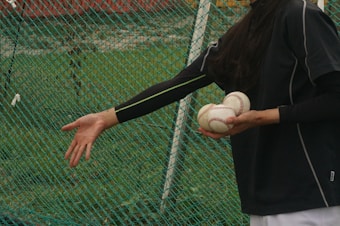 A person dressed in dark sportswear holds multiple baseballs in one hand while reaching out with the other hand. The background consists of a green mesh net and grassy area, suggesting a sports field or training area.