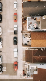An aerial view of a residential area showing houses with their rooftops and a section of a street. On the street, there are multiple parked cars positioned along the sidewalk. The top view also reveals some courtyards and patios with outdoor furniture such as chairs and tables. The area appears to be an organized urban setting.