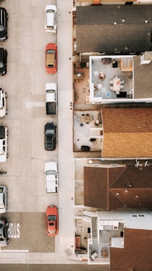 An aerial view of a residential area showing houses with their rooftops and a section of a street. On the street, there are multiple parked cars positioned along the sidewalk. The top view also reveals some courtyards and patios with outdoor furniture such as chairs and tables. The area appears to be an organized urban setting.