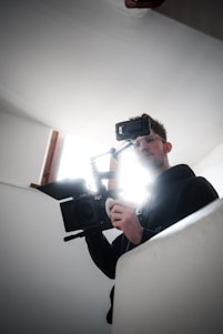 A person is holding a professional video camera, possibly in the process of filming. The light source behind them creates a halo effect around their head, casting a dramatic silhouette. The setting appears to be indoors with plain white walls.