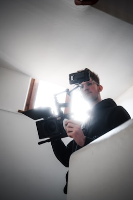 A person is holding a professional video camera, possibly in the process of filming. The light source behind them creates a halo effect around their head, casting a dramatic silhouette. The setting appears to be indoors with plain white walls.