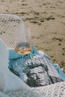 A wine glass with a light-colored beverage sits atop a magazine featuring a black and white portrait. The scene is set on a beach, with a blanket and a pair of sunglasses casually placed nearby. Shadows from a net-like fabric create patterns on the items.