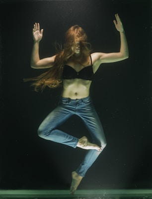 A woman is submerged underwater, wearing a black bra and blue jeans. Her long hair flows around her, and she holds her arms up with palms open. Her legs are crossed in a pose that suggests weightlessness.