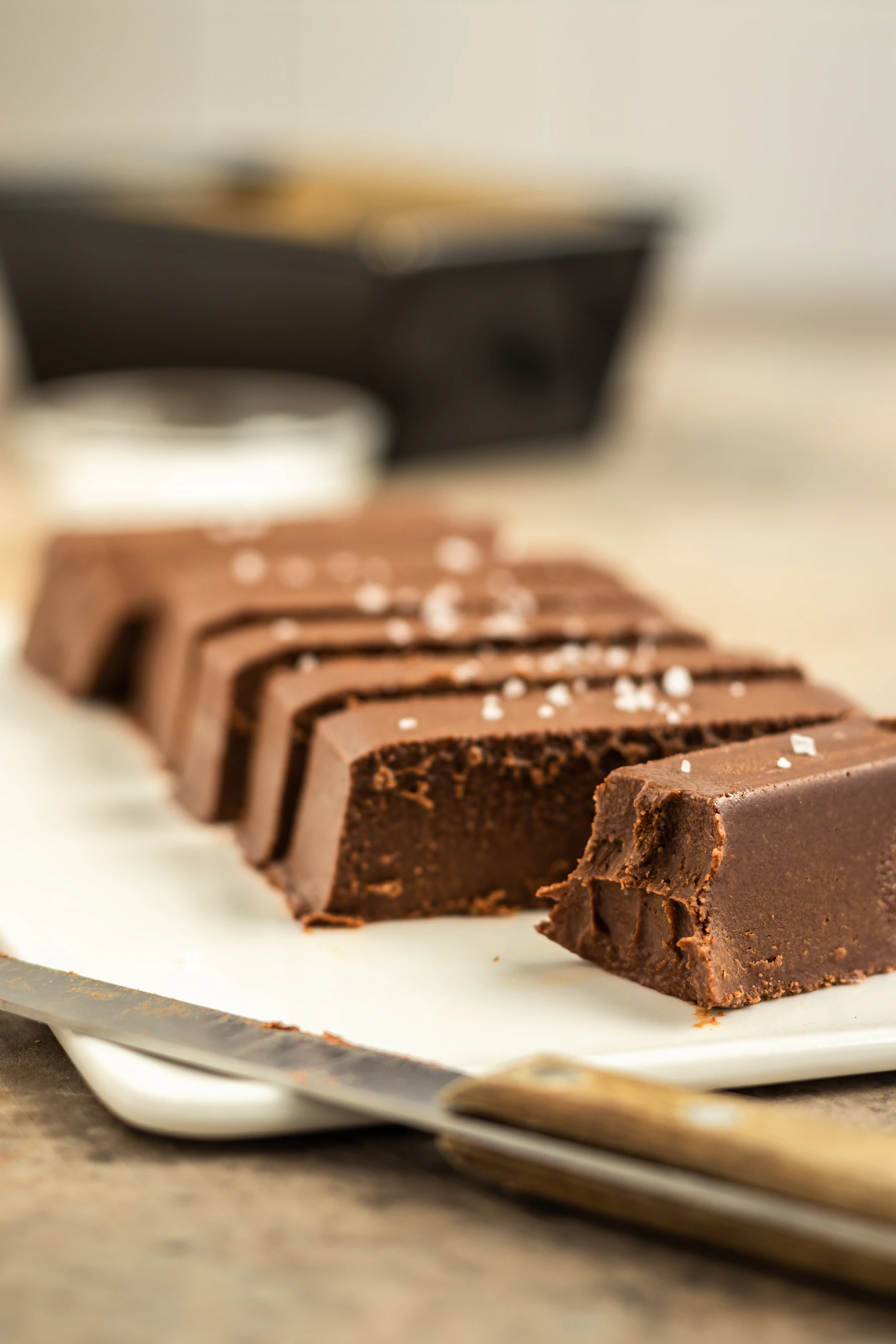 A bar of chocolate fudge cut into slices and topped with flaky salt, a knife in front