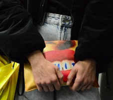 A person holds a colorful clutch bag against their denim jeans. The bag features a vivid design with bright red lips and blue highlights on a yellow and orange background. The person is wearing a dark jacket, and there's a yellow plastic bag partially visible.