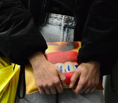 A person holds a colorful clutch bag against their denim jeans. The bag features a vivid design with bright red lips and blue highlights on a yellow and orange background. The person is wearing a dark jacket, and there's a yellow plastic bag partially visible.