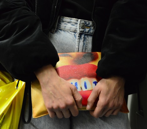 A person holds a colorful clutch bag against their denim jeans. The bag features a vivid design with bright red lips and blue highlights on a yellow and orange background. The person is wearing a dark jacket, and there's a yellow plastic bag partially visible.