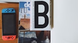 A combination of a Nintendo Switch with blue and red controllers placed next to a stack of magazines with prominent text and images. The top magazine has a large 'B' on its cover, and another displays an architectural design.