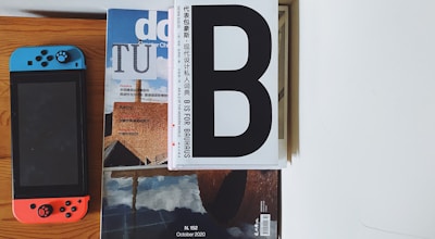 A combination of a Nintendo Switch with blue and red controllers placed next to a stack of magazines with prominent text and images. The top magazine has a large 'B' on its cover, and another displays an architectural design.