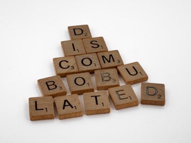 Wooden Scrabble tiles are arranged to spell out the word 'DISCOMBOBULATED' in the shape of a triangle on a white background. Each tile has a letter with an associated point value displayed in the bottom right corner.
