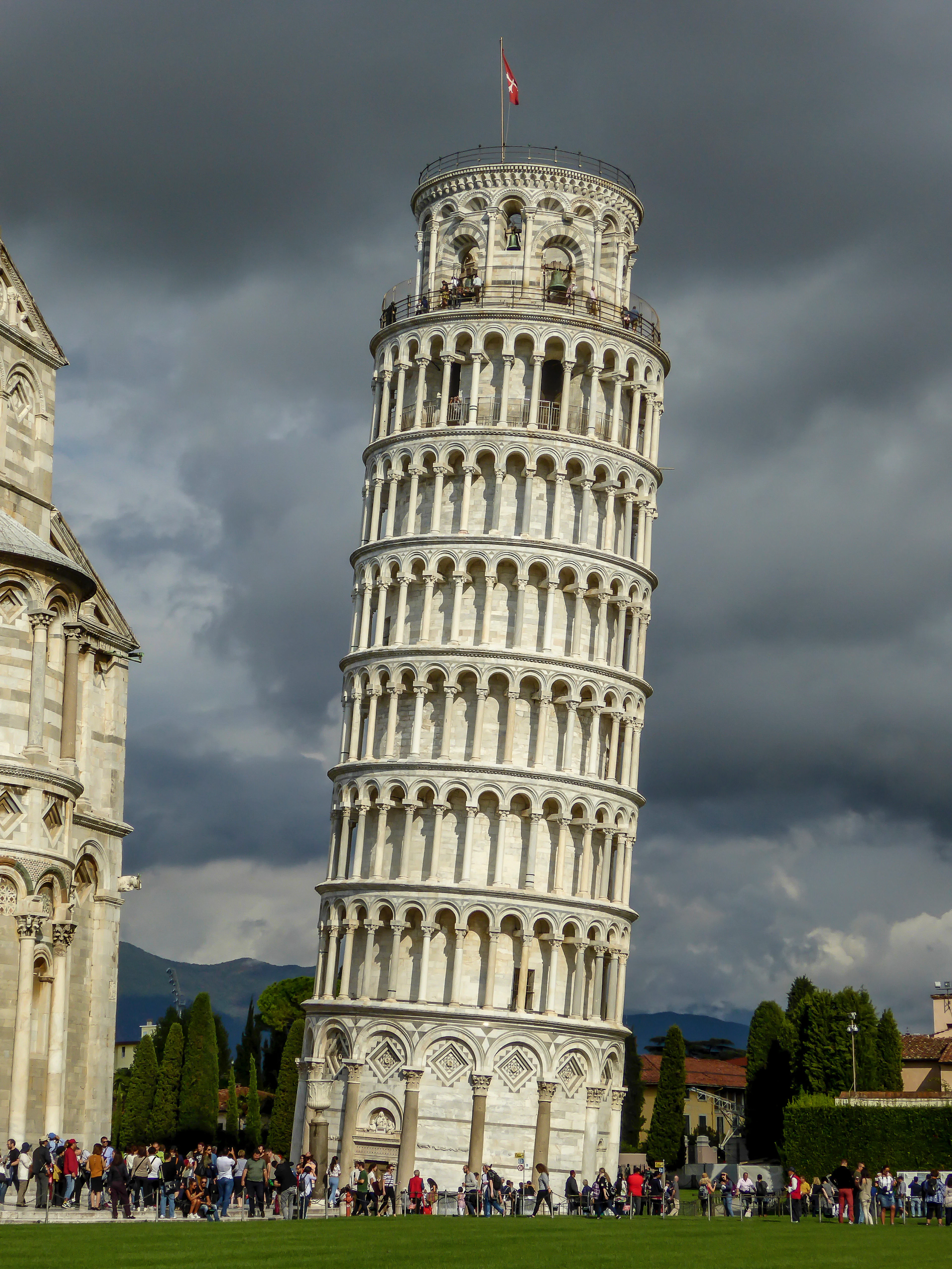 The Leaning Tower stands white against dark storm clouds, a crowd gathered at its base.