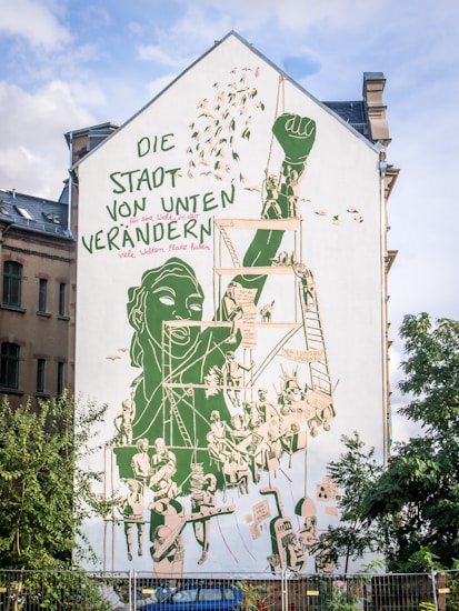A large mural is painted on the side of a building, featuring a dominant green and beige color scheme. The artwork depicts a group of people engaged in various activities, including climbing a ladder and raising a fist, symbolizing resistance and community action. There's text in German integrated into the design, which emphasizes change and collaboration.