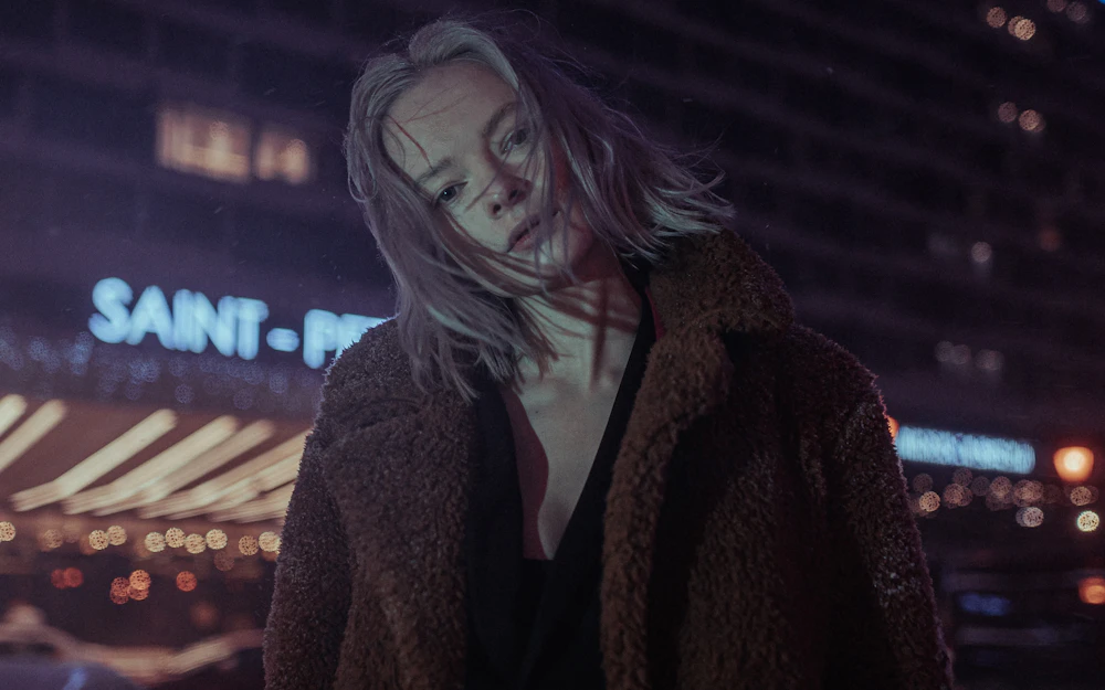 A woman in a brown coat beside a shopfront, photographed well after closing time