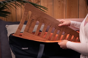 A person is holding a wooden board with slots, which has 'Harmoni' engraved on it. They are placing it into a black bag with an orange interior lining. The setting includes a neutral-colored couch and a wooden paneled wall, with a green plant visible to the side.