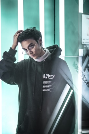 A person with short dark hair and glasses is wearing a black NASA hoodie. The background features bright vertical white lights against a teal-colored wall, giving a futuristic and modern aesthetic.