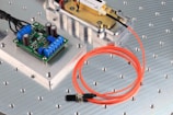 The image features an electronic circuit board placed on a metallic surface with perforated holes. Blue terminal connectors and cylindrical capacitors are visible on the green board. Nearby, an orange fiber optic cable is coiled beside a metal component encased in a transparent cover. The setting appears to be a technical or engineering workspace.