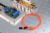 The image features an electronic circuit board placed on a metallic surface with perforated holes. Blue terminal connectors and cylindrical capacitors are visible on the green board. Nearby, an orange fiber optic cable is coiled beside a metal component encased in a transparent cover. The setting appears to be a technical or engineering workspace.