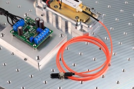 The image features an electronic circuit board placed on a metallic surface with perforated holes. Blue terminal connectors and cylindrical capacitors are visible on the green board. Nearby, an orange fiber optic cable is coiled beside a metal component encased in a transparent cover. The setting appears to be a technical or engineering workspace.
