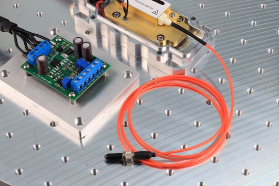 The image features an electronic circuit board placed on a metallic surface with perforated holes. Blue terminal connectors and cylindrical capacitors are visible on the green board. Nearby, an orange fiber optic cable is coiled beside a metal component encased in a transparent cover. The setting appears to be a technical or engineering workspace.
