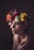 A person is adorned with a vibrant floral crown composed of various flowers in shades of red, white, yellow, and green. The lighting is soft and moody, casting gentle shadows across the face, conveying a contemplative or serene expression.