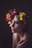 A person is adorned with a vibrant floral crown composed of various flowers in shades of red, white, yellow, and green. The lighting is soft and moody, casting gentle shadows across the face, conveying a contemplative or serene expression.